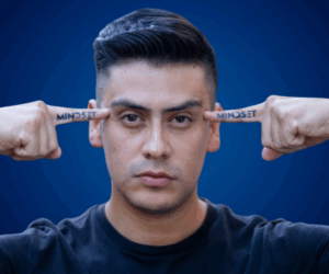 Retrato de Manuel Vásquez Arce apuntando con los dedos a sus sienes, donde tiene tatuada la palabra "MINDSET", con fondo azul y expresión decidida.