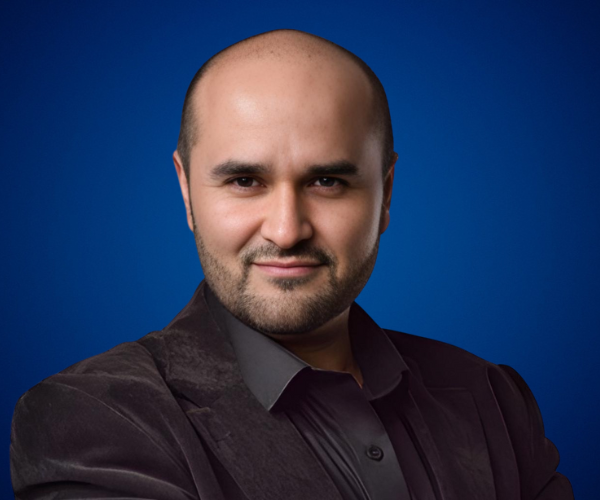 Fotografía de Fernando Veliz posando con confianza frente a un fondo azul, luciendo saco oscuro y camisa negra.